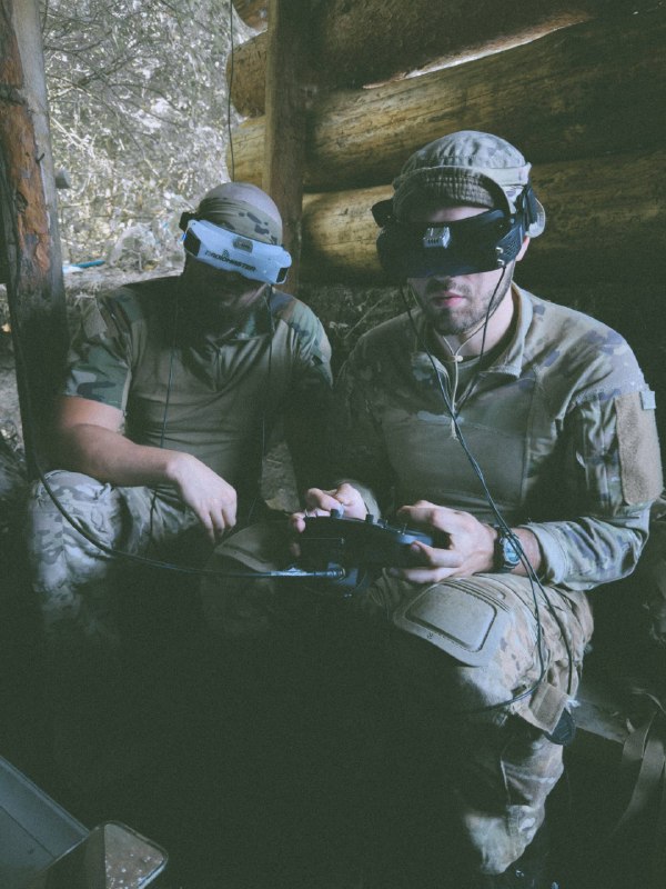 Two operators in a covered field position preparing and flying an FPV system with goggles and a controller